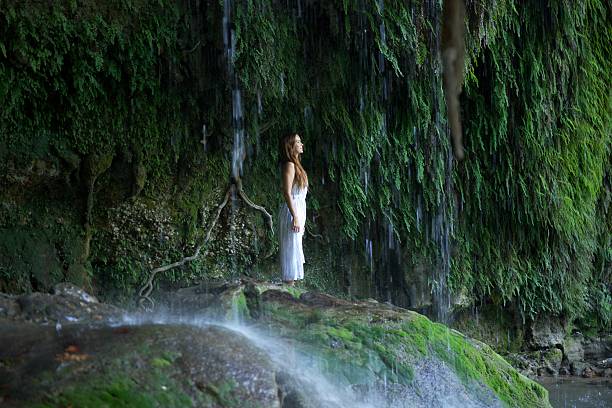 A peaceful woman in a white dress stands beneath the cascading Antalya Famous Duden Waterfall, surrounded by lush green moss, flowing water, and serene wilderness in a calming natural setting.
