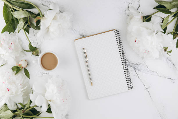 Beautiful white peony flowers with empty notebook on white marble stone background, copy space for your text or design, top view, flat lay