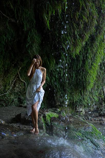 A woman in a white dress stands near a cascading waterfall, touching her hair as she enjoys the refreshing mist, surrounded by lush greenery and moss-covered rocks.