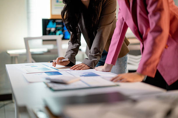 istock Professional women analyzing financial charts, gesturing with pen over conference table in modern, sunlit workspace 2203939297