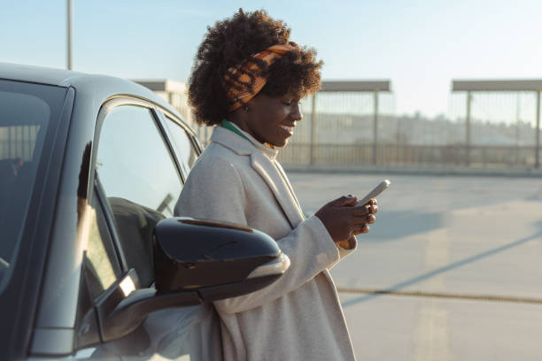 istock Smiling woman using smartphone while leaning against a car in the sun 2203950277