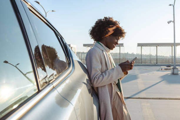 istock Woman smiling outdoors while holding smartphone and standing near a car 2203950430