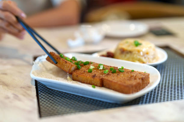 A close-up of crispy fried turnip cake garnished with green onions, served on a white plate, with a hand using chopsticks to pick up a piece in a restaurant setting.