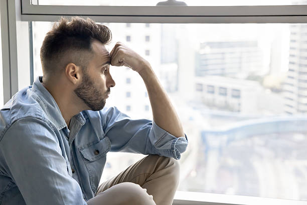 istock Upset man seated on windowsill looks out window 2203958875