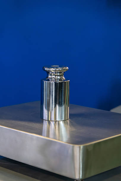 A shiny metal weight stands upright on a reflective silver platform. The setting features a smooth surface and a solid blue background, suggesting a laboratory or measurement context.