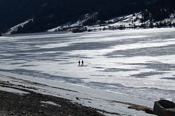 Two people walk across a vast frozen lake, surrounded by a serene winter landscape with snow-covered mountains and scattered houses in the background. The icy surface glistens under the cold sunlight, creating a tranquil yet adventurous scene.