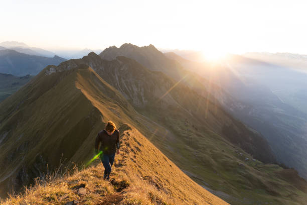Young man hikes along narrow trail on top of high mountain ridge at sunrise, sun rises over distant mountains, Valais, Swiss Alps