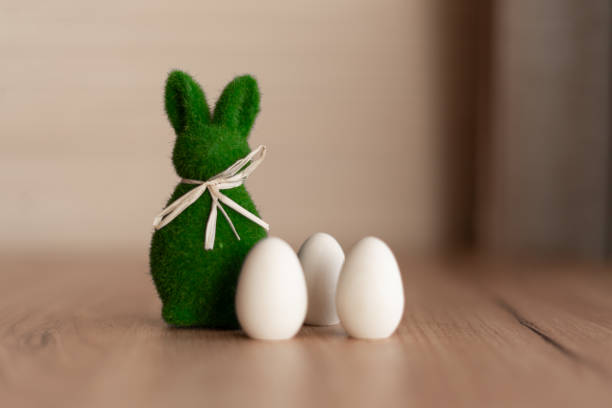 A green Easter bunny figurine with a rustic ribbon stands beside three white wooden eggs on a wooden surface. The soft lighting and neutral tones create a cozy, minimalist holiday atmosphere.