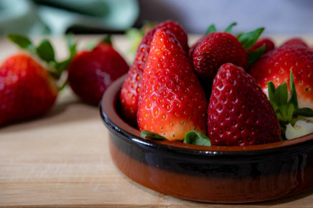 A bowl of strawberries sits on a wooden table. The bowl is filled with a variety of strawberries, some of which are larger and others smaller