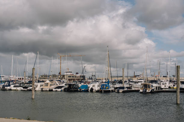 A serene marina filled with yachts and sailboats docked under a dramatic overcast sky. The calm water reflects the moody atmosphere, while a crane and waterfront buildings in the background add an industrial contrast to this coastal scene. Perfect for projects related to boating, travel, or maritime lifestyles.