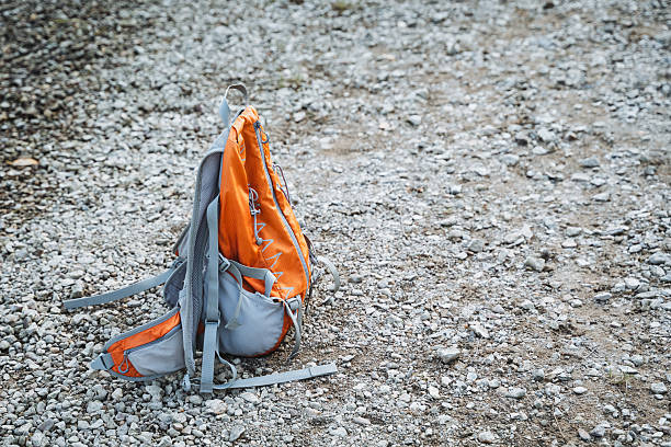 An orange and gray backpack is currently laying on the ground, resting on the surface of the earth, surrounded by various elements such as soil, rocks, and possibly some litter nearby