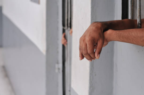istock prisoner's hands behind bars with black background 2203988424