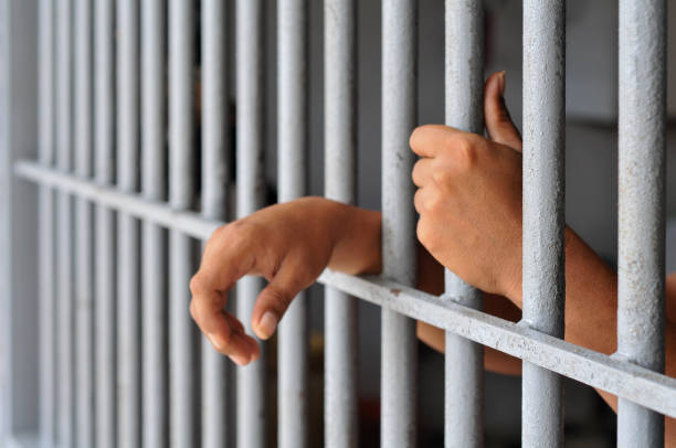istock prisoner's hands behind bars with black background 2203989504