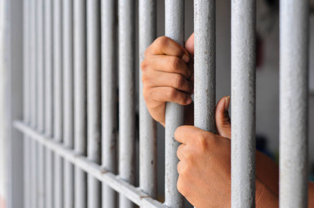 istock prisoner's hands behind bars with black background 2203989508