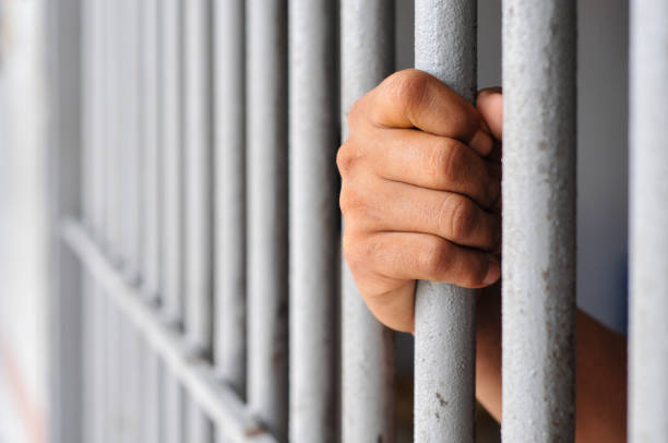 istock prisoner's hands behind bars with black background 2203989553