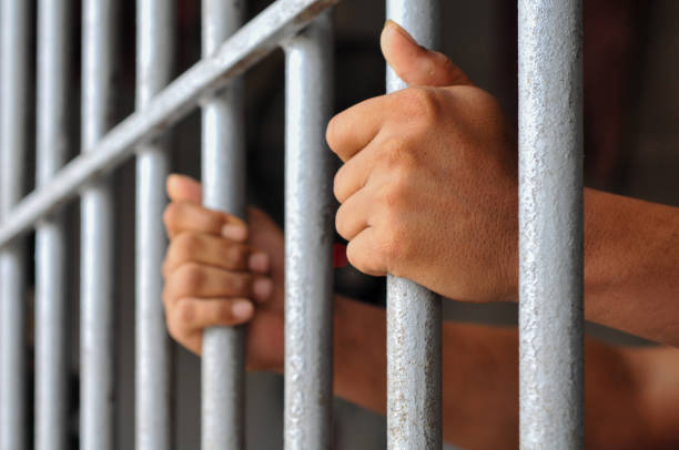 istock prisoner's hands behind bars with black background 2203989591