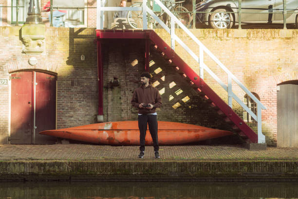 Young man holding a drone remote controller standing next to a canoe in a dutch canal in a sunny day