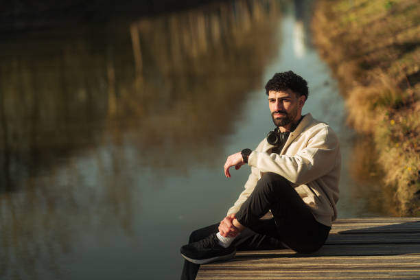 Young man sitting on a wooden pier by the river, enjoying the sunset after exercising