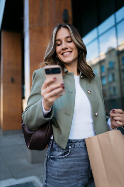 istock Smiling young woman using smartphone while carrying shopping bag 2204028238