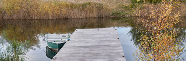 A peaceful scene featuring a wooden dock and a small boat on a tranquil lake. The surrounding autumn landscape showcases golden reeds and sparse trees, enhancing the serene atmosphere. This picturesque setting, often found in Denmark, captures the essence of quiet nature and a calm retreat.