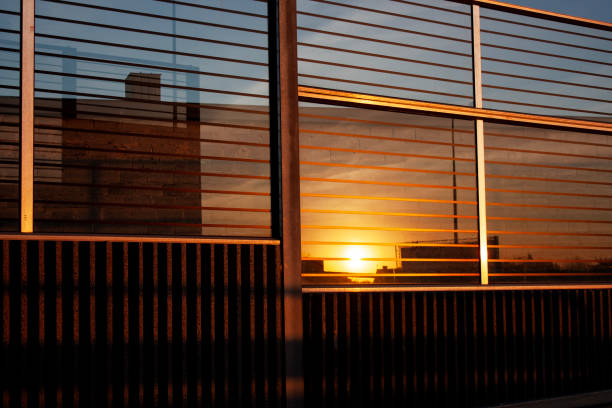 A warm, golden sunset reflecting off the sleek metal and glass facade of a modern urban structure. The interplay of light and shadows creates a geometric abstract composition, highlighting architectural minimalism and industrial aesthetics. This atmospheric image conveys themes of urban lifestyle, modern design, and the beauty of golden hour reflections.