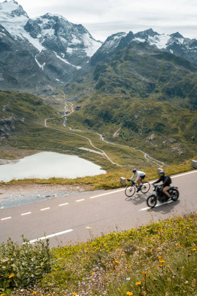 Road biker passes motorcycle on ride up high mountain pass, lake below, Swiss Alps