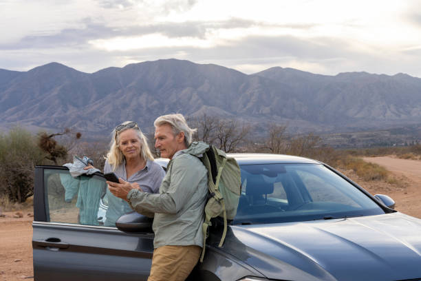istock Mature couple lean against car and use mobile phone 2204042159