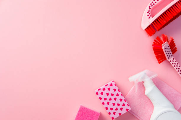 Cleaning supplies on a pink background, featuring brushes, a spray bottle, and heart-patterned sponges. Ideal for Valentine's Day cleaning themes or romantic household chores
