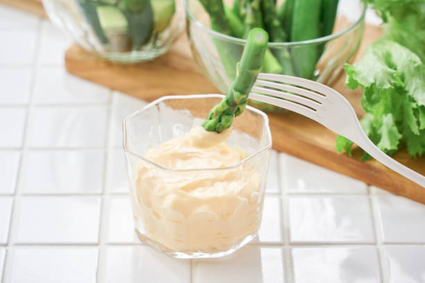 A close-up shot of mayonnaise in a small glass bowl. Fresh lettuce, asparagus, and snap peas are placed in the background, creating a fresh and simple composition. This image showcases a creamy and appetizing mayonnaise.