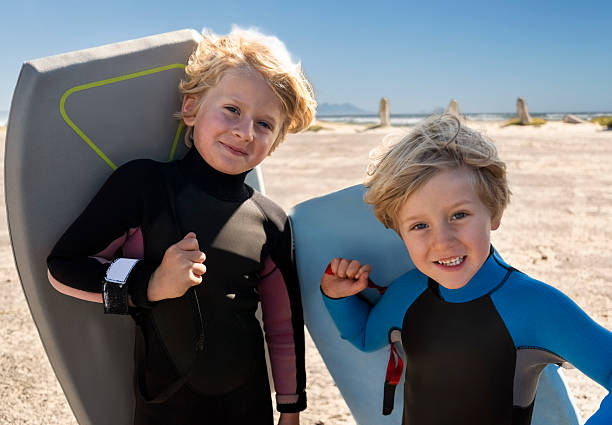 Portrait of small boys dressed in wetsuits carrying their surfboards and standing on the beach