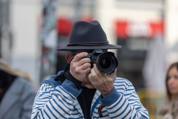 Capturing the Moment in Plaza de Callao, Madrid, Spain. A street photographer in a fedora and striped jacket focuses intently through his camera lens in Plaza de Callao, one of Madrid’s most vibrant squares. Surrounded by the energy of the city, he captures the urban landscape, blending into the dynamic rhythm of daily life. The blurred background hints at the bustling activity of this iconic location, where culture, commerce, and creativity converge.
