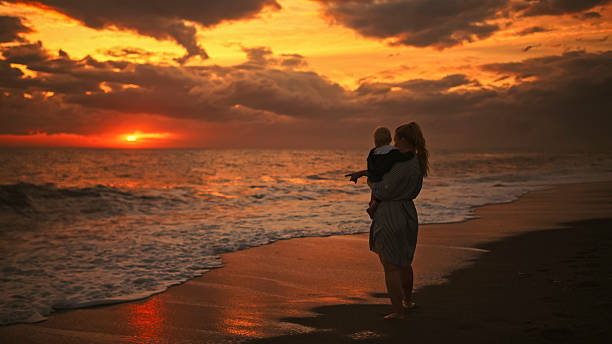 Mother Gently holds her Child in her Arms as they Stand at the Edge of the Sea During a Breathtaking Sunset,The Woman Points Toward the Glowing Horizon,Sharing a Moment of Wonder and Connection Beneath the Dramatic Evening Sky