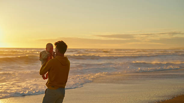 istock Father Holding Baby at the Beach During Serene Sunset 2204120681