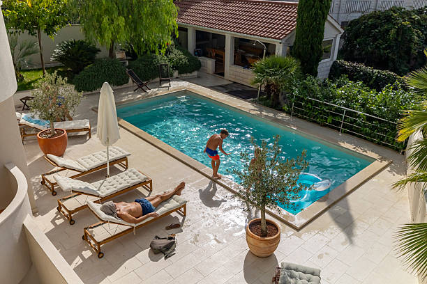 Aerial view of young men in swim shorts lounging and swimming in private pool area. Mediterranean-style villa features cream-tiled deck, cushioned sunbeds, potted olive trees, and lush garden landscaping.