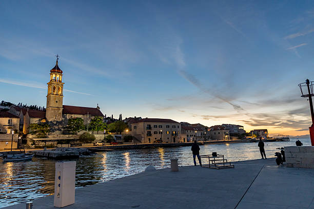 A peaceful evening in Sutivan, Brac?, with a softly lit historic church, calm harbor waters, and people strolling along the waterfront at sunset.