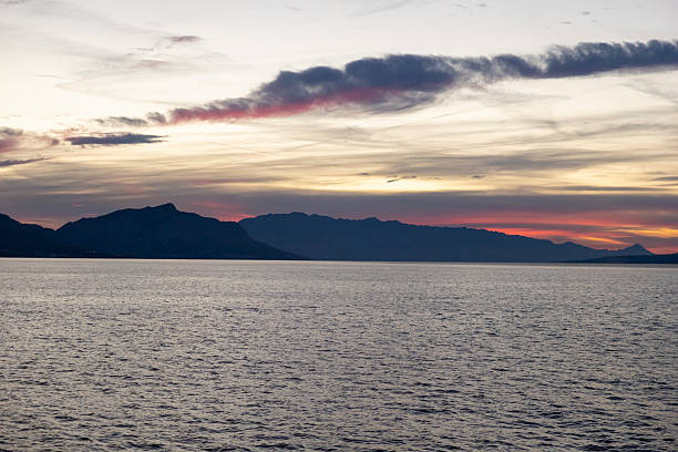 A tranquil sunset over Croatian islands featuring a colorful sky blending orange, pink, and violet hues, calm Adriatic waters, and silhouetted mountains on the horizon. The peaceful setting evokes relaxation and natural beauty.