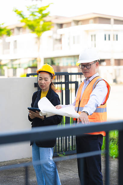 istock Two male and female architectural engineers are looking at a building with laptops in front. They are wearing safety equipment and pointing at something on a building. Vertical image 2204153339