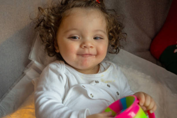 A cheerful toddler with curly hair and a bright smile is lying down while holding a colorful cup. The child’s happy expression and relaxed posture create a warm and joyful moment in a cozy home setting