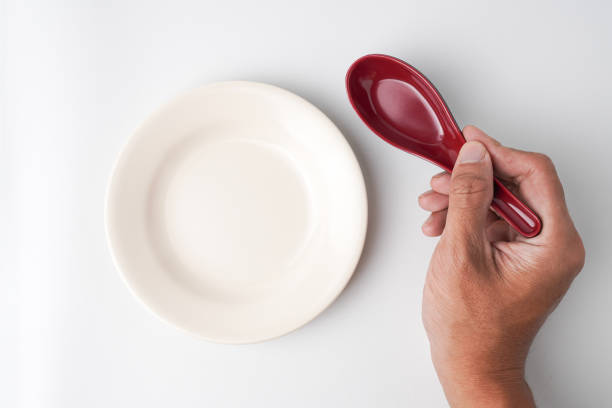 Someone hand is holding red colored chinese soup spoon next to empty beige colored round shaped plate. Isolated on white background. Flat Lay Setup