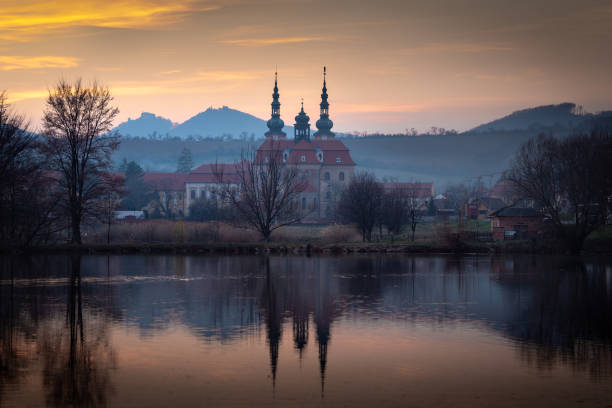 The image depicts a church by a serene lake at sunset, surrounded by trees and hills, with a peaceful and tranquil atmosphere, showcasing a picturesque countryside setting with reflective water.