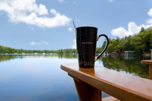 A coffee mug sits on the arm of an Adirondack chair, facing a calm blue lake. Cottage cabins line the shore, nestled among green trees, creating a peaceful and cozy lakeside scene. Copy space.