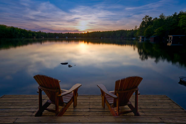 Two Adirondack chairs sit on a wooden dock at sunset, overlooking a tranquil Muskoka lake. The water glows in the fading light, and nearby cabins dot the shore, capturing the essence of summer.
