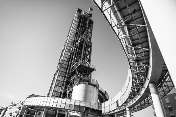A wide-angle black and white image of an oil refinery with a towering distillation column and curved pipeline structures. Industrial metal framework contrasts against a clear sky