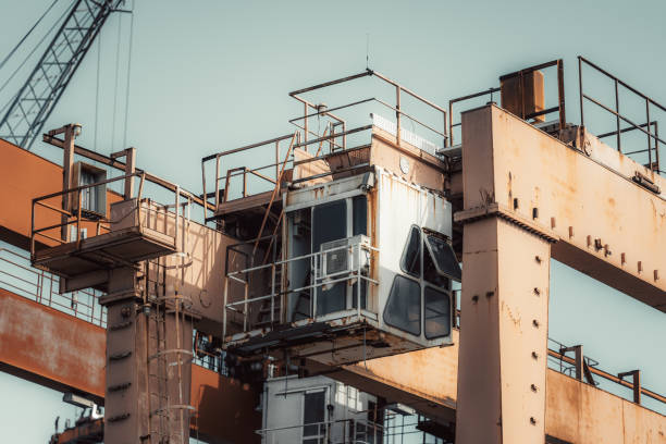 Close-up telephoto shot of an aged industrial crane control cabin with rusted metal surfaces, ladders, and railings. The structure shows signs of wear, set against a clear sky background