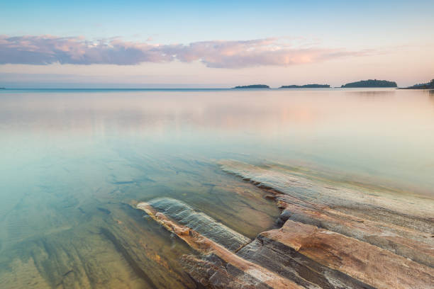Water reflects soft hues of twilight at Lake Vanern in Sweden, while smooth rocks frame the serene shoreline. Gentle ripples create a peaceful atmosphere during dusk.