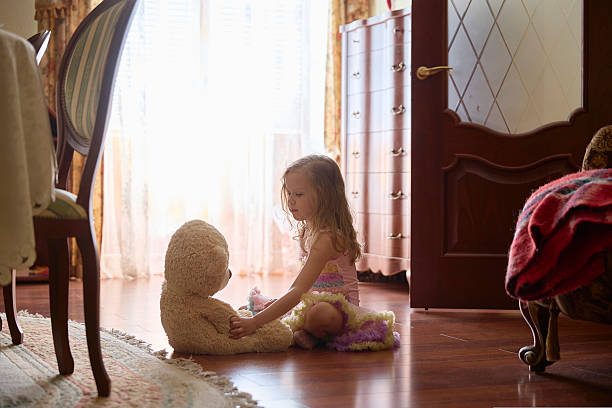 A young girl sits on the wooden floor, gently interacting with a soft teddy bear in a warmly lit room filled with sunlight. Cozy furniture surrounds them, creating a homey atmosphere.