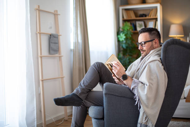 Profile view of smiling man reading book sitting in armchair wrapped in blanket.