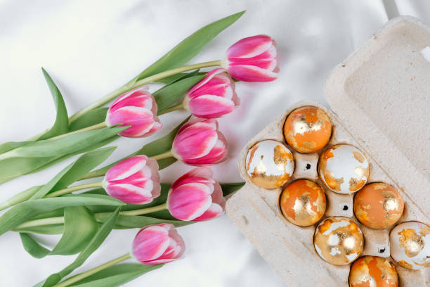 istock Painted gold Easter eggs in a carton on a white silky background with pink tulips 2204203199
