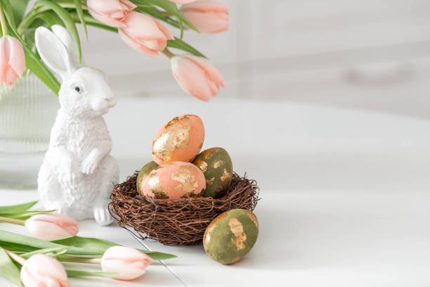 istock A bouquet of delicate tulips, Easter bunnies and eggs with a golden pattern on the table. There is a white kitchen in the background. Beautiful greeting card. The concept of minimalism. 2204203881