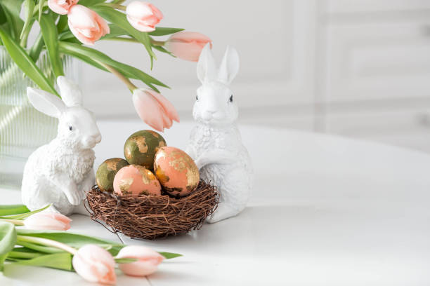 istock A bouquet of delicate tulips, Easter bunnies and eggs with a golden pattern on the table. There is a white kitchen in the background. Beautiful greeting card. The concept of minimalism. 2204203882